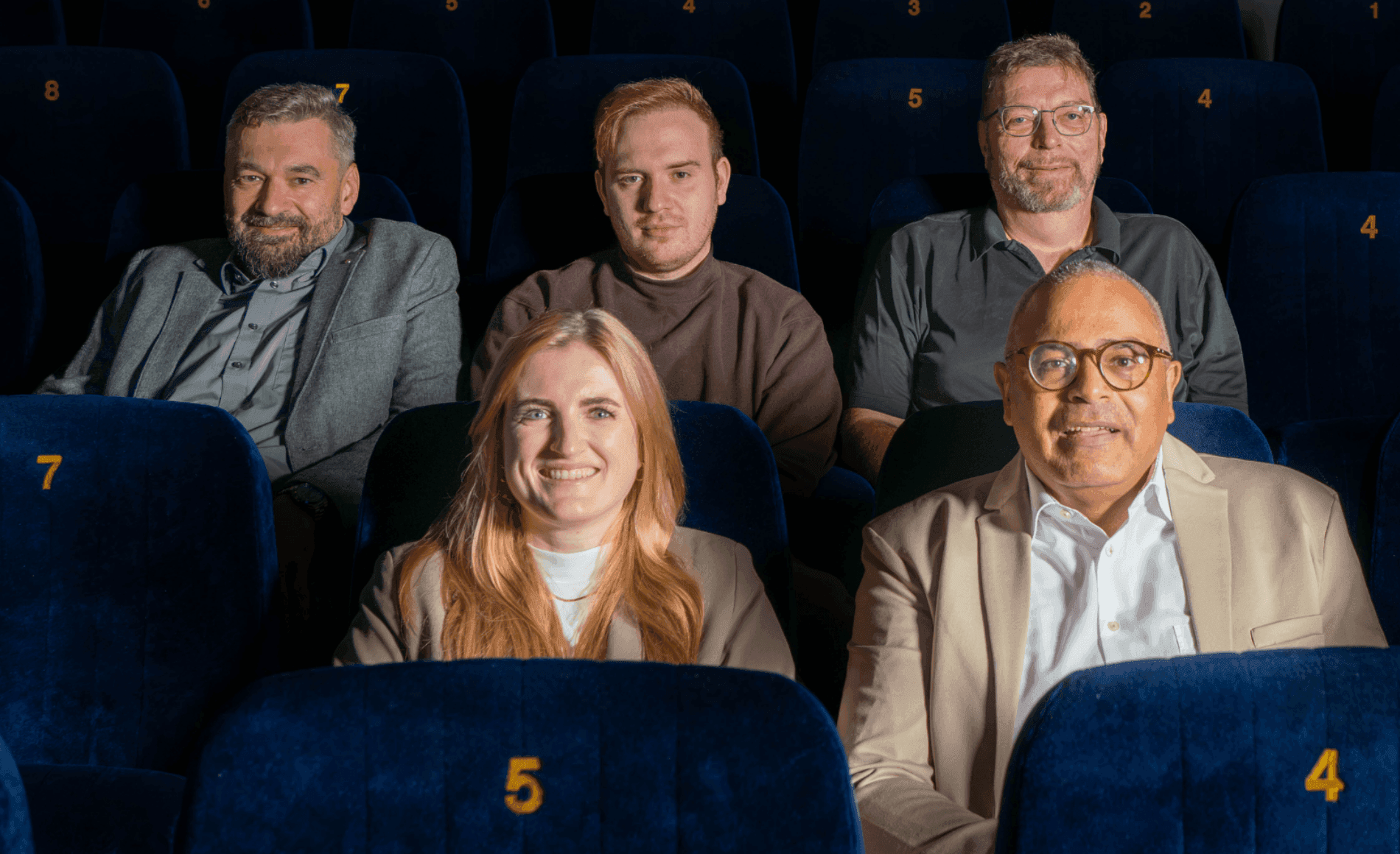 Four men and one woman smiling, seated in dark blue movie theater seats.