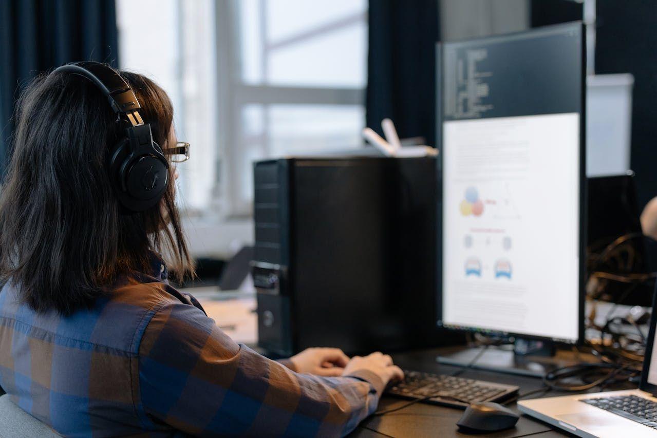 Person wearing headphones intently working on a dual-monitor computer setup, one vertical.